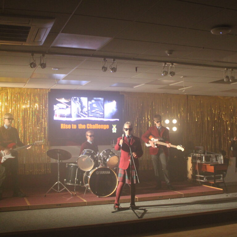 School band on stage with gold fringe backdrop; screen behind reads 'Rise to the Challenge'.